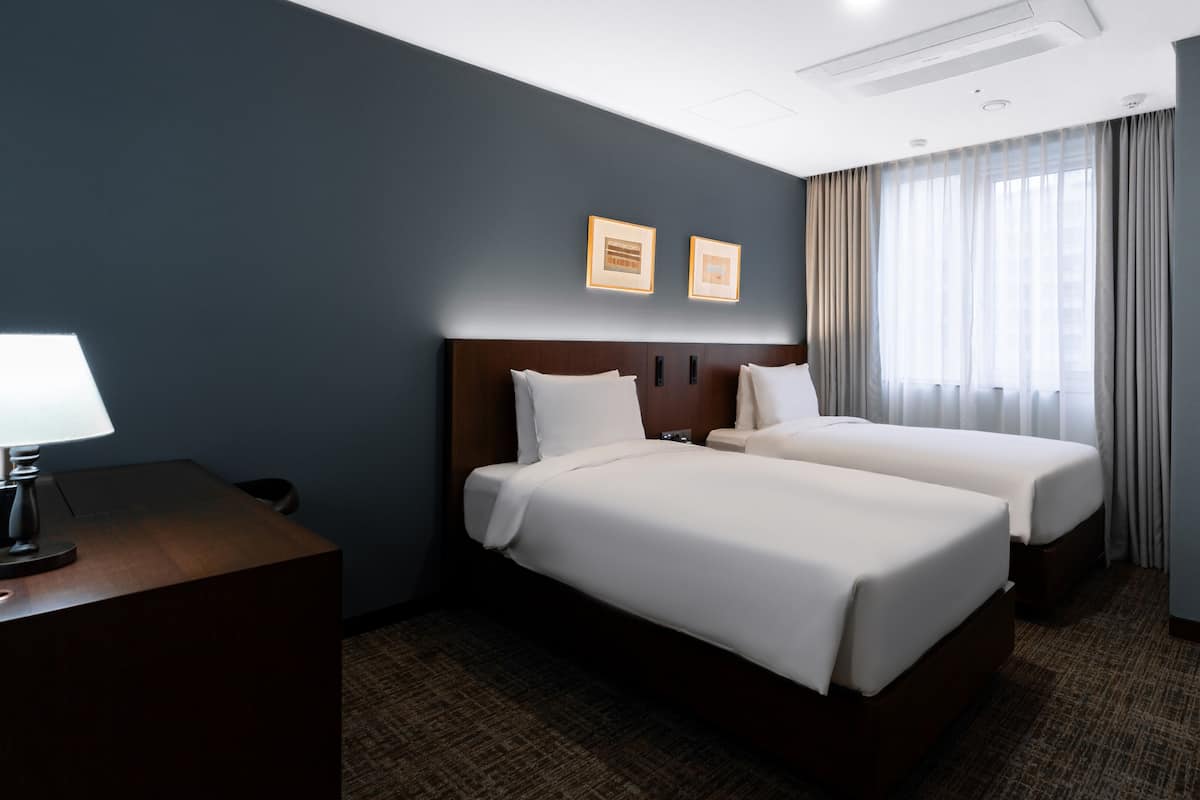 Une chambre d’hôtel avec deux lits, un bureau avec une lampe, une chaise et une fenêtre avec des rideaux.