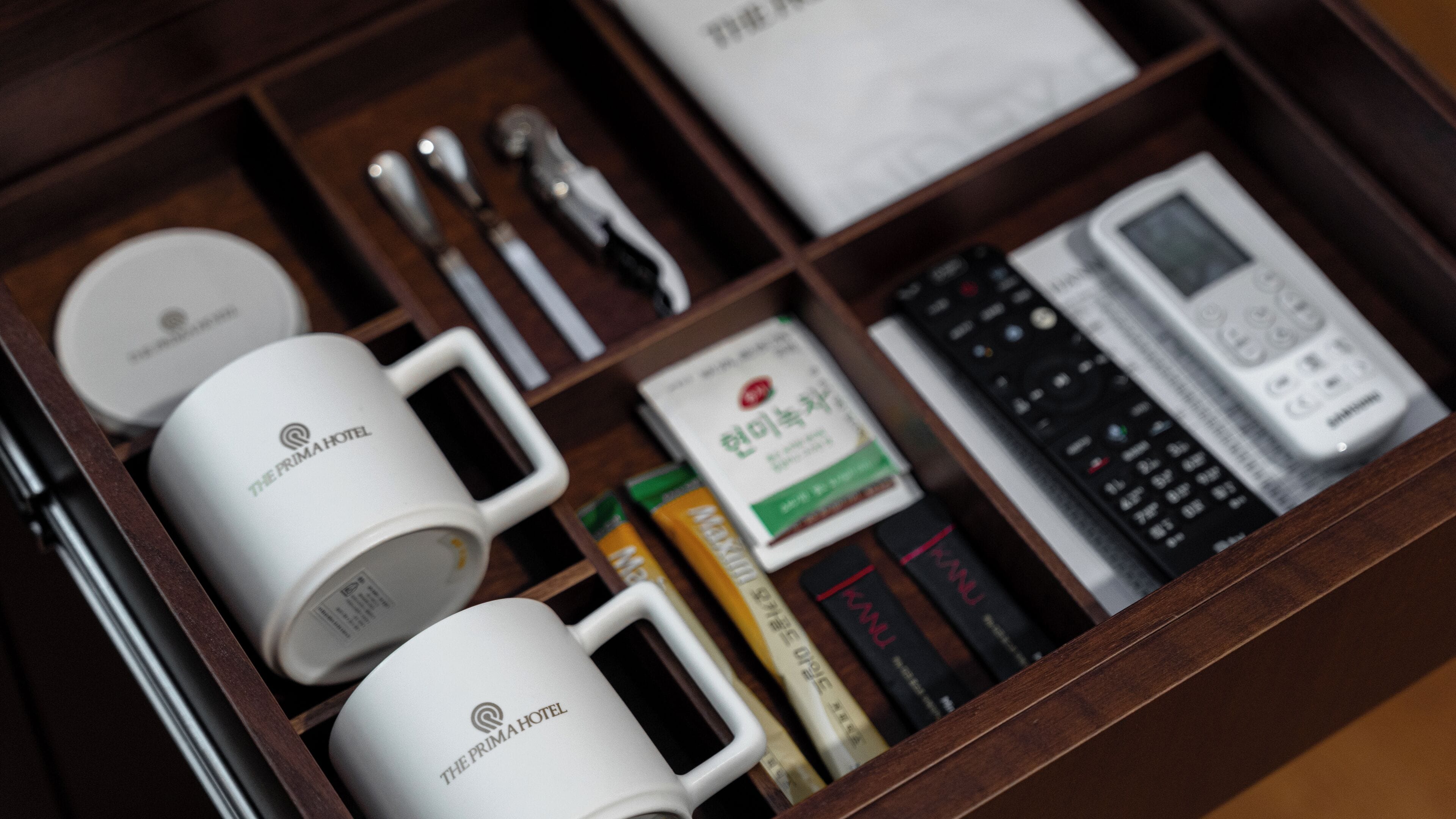 A wooden tray with a white mug labeled "The Prima Hotel," a remote control, a book, and a small box of tissues.