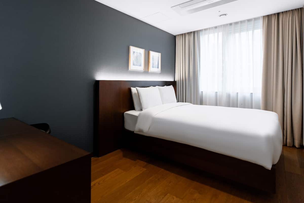 Une chambre d’hôtel avec un bureau en bois, un lit recouvert de draps blancs, un mur sombre et une fenêtre avec des rideaux.