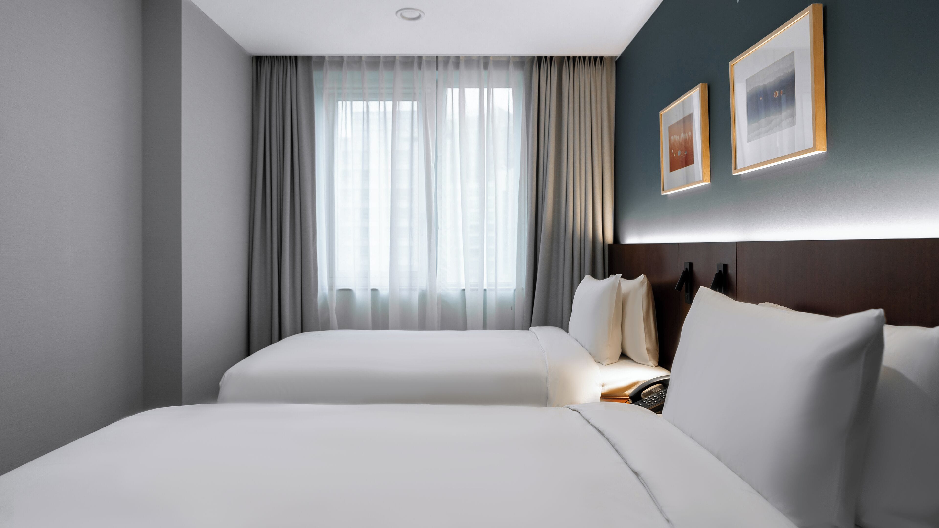 Habitación de hotel con dos camas, un ventanal grande con cortinas traslúcidas, un cabecero oscuro y dos cuadros en la pared.