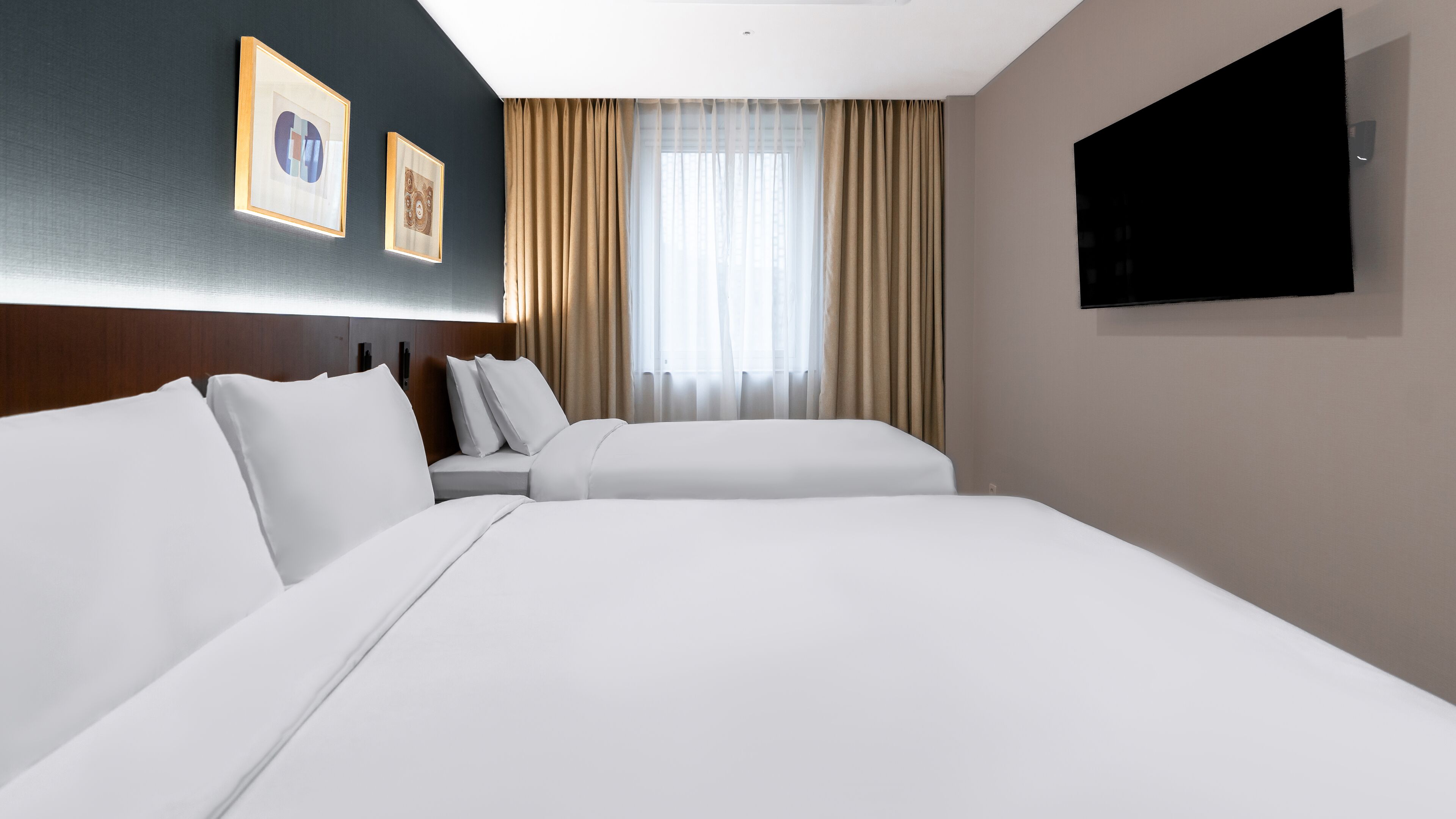 Une chambre d’hôtel avec deux lits, une grande fenêtre avec des rideaux, un téléviseur à écran plat fixé au mur et des œuvres d’art encadrées accrochées au mur.