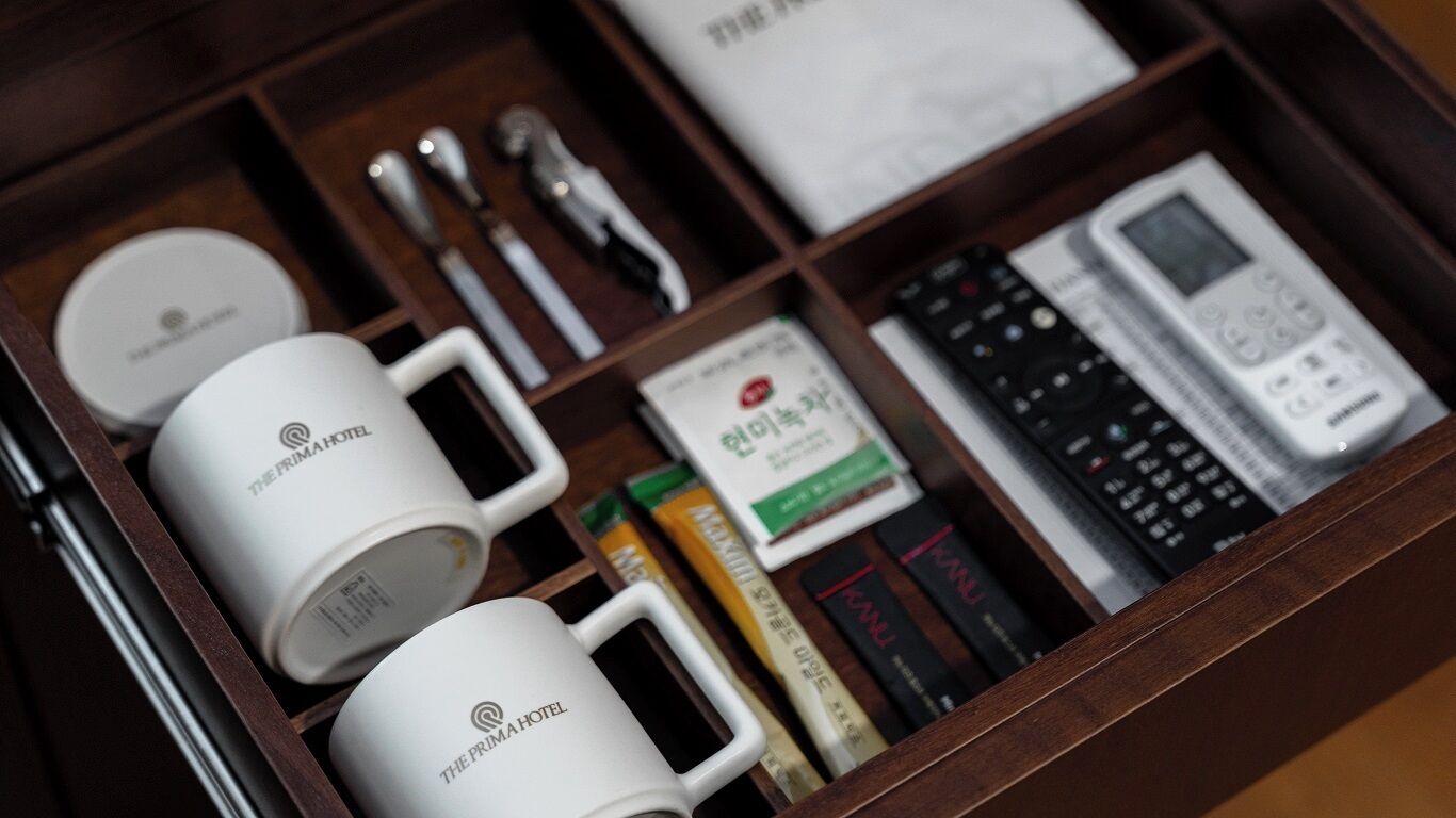 A drawer with white mugs labeled "The Prima Hotel," a remote control, a book, and a box of tissues.