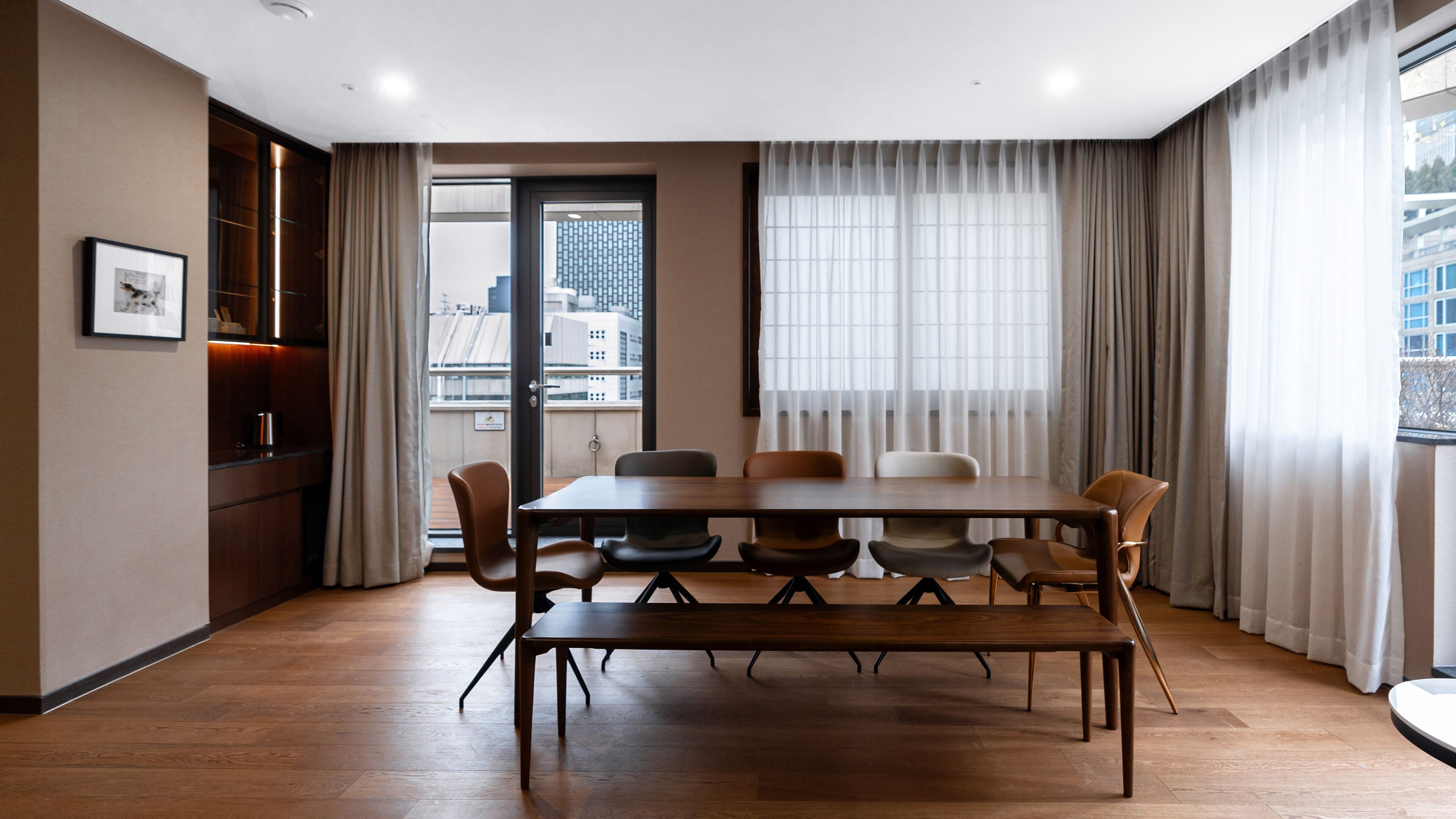 Una sala de conferencias moderna con una mesa y sillas de madera, ventanas amplias con cortinas y vistas a la ciudad.