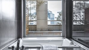 A modern bathroom with a large bathtub, a window with a view of buildings and trees, and a wooden panel on one wall. - The Prima Hotel Jongno Insadong (Seoul)