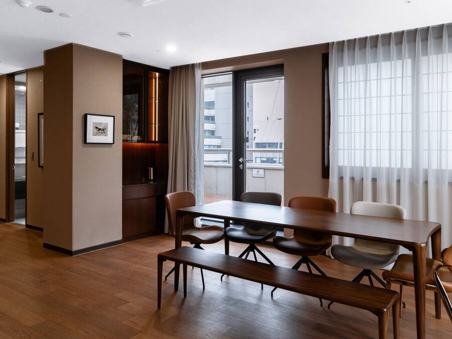 Quarto de hotel com piso de madeira, mesa de jantar com cadeiras, cama e uma grande janela com cortinas.