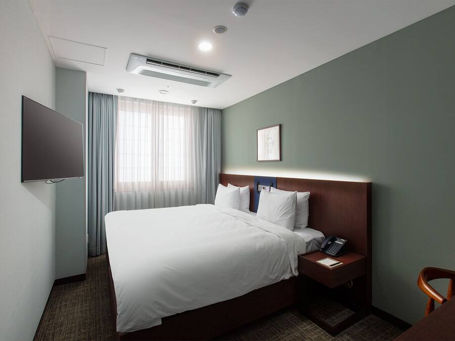 A hotel room with a bed, a bedside table with a phone and a notepad, a wall-mounted TV, and an air conditioning unit.