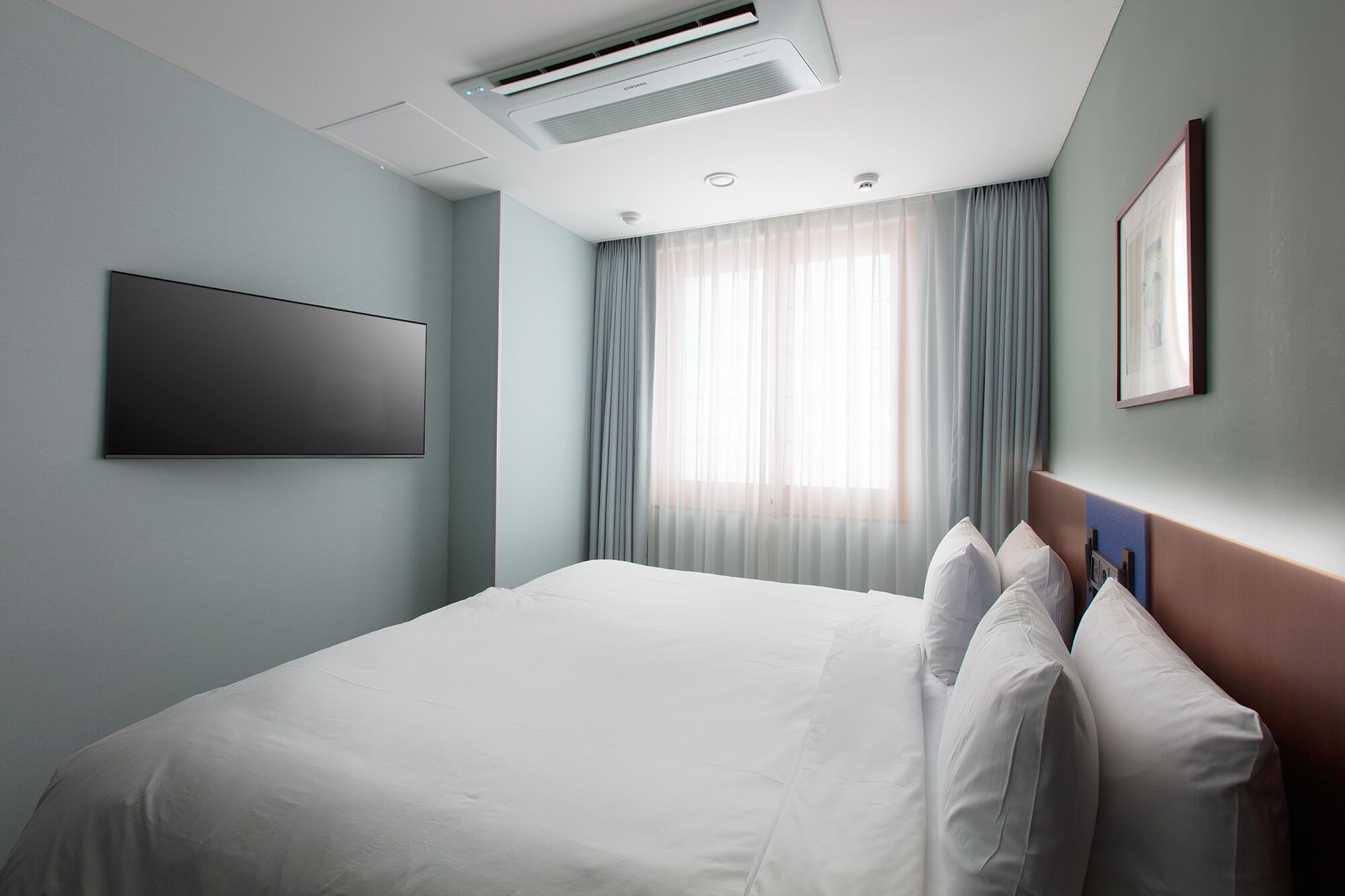 A modern hotel room with a large bed, a flat-screen TV mounted on the wall, and a window with sheer curtains.