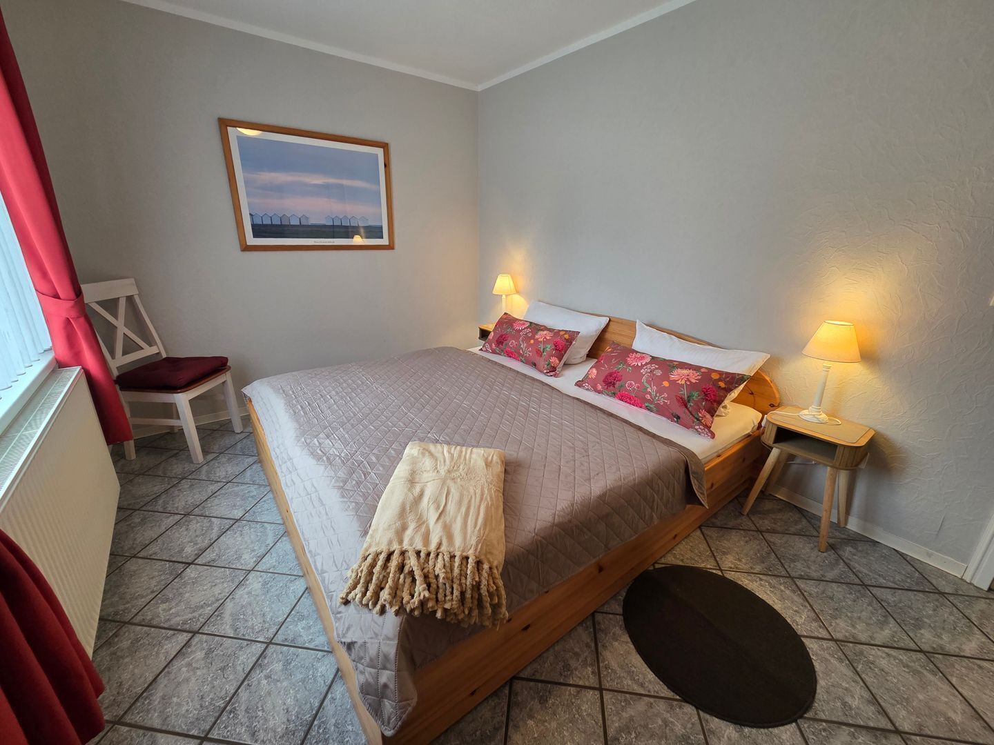 Een slaapkamer met een bed, twee bedlampjes, een stoel, een klein tafeltje, een foto aan de muur en een raam met gordijnen.