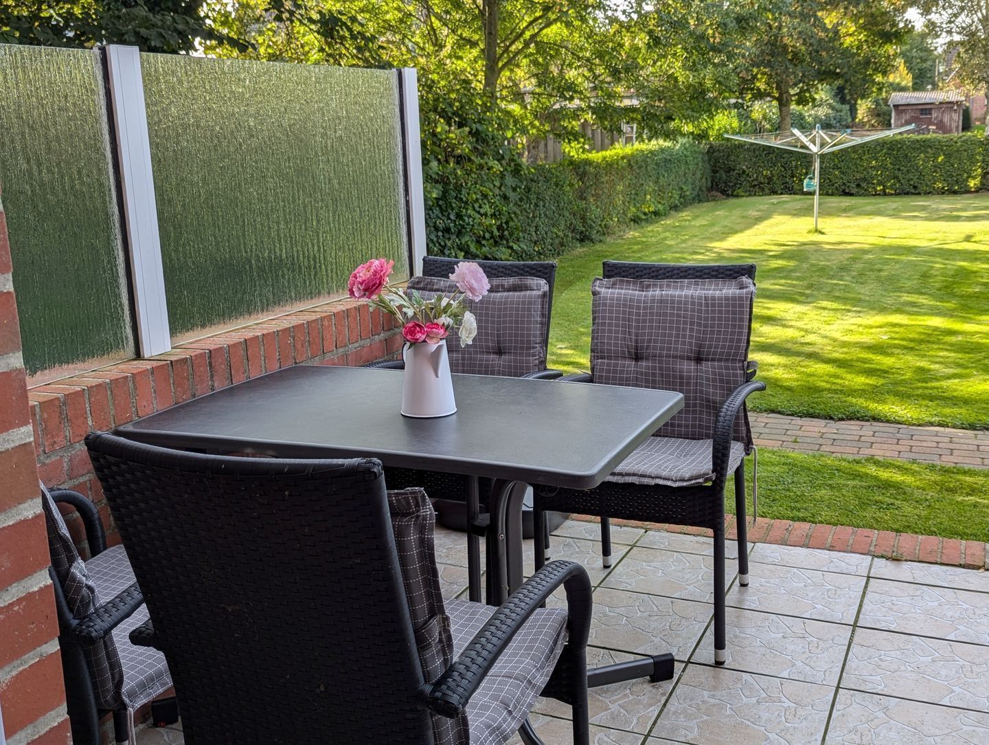 Un patio exterior con una mesa y sillas, un jarrón blanco con flores y un césped bien cuidado.