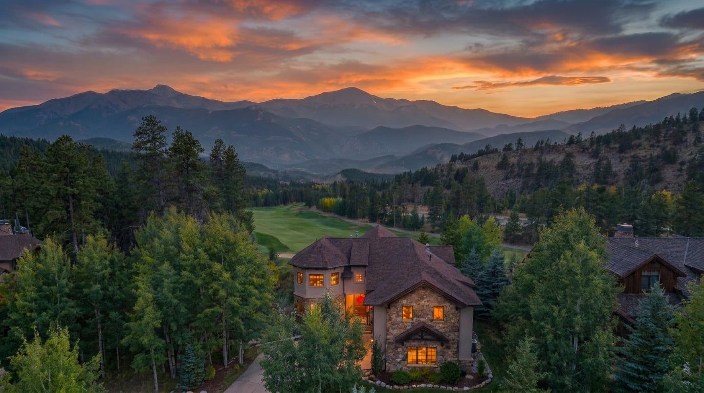 Une grande maison à la façade en pierre, dotée d’une tourelle, entourée d’une forêt dense et avec des montagnes en arrière-plan.