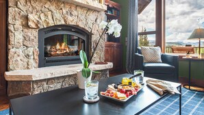 Una chimenea de piedra con fuego, una mesa con una tabla de embutidos, una orquídea en maceta y vistas al exterior.