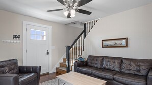 A living room with a dark brown leather sofa, a light wooden coffee table, and a ceiling fan. A staircase with white railings leads to another level. - Steps to Hampton Beach and all entertainment! 4 bedrooms, pet friendly! (Hampton)