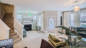 A spacious living room with a fireplace, a staircase, and a large window with blinds.