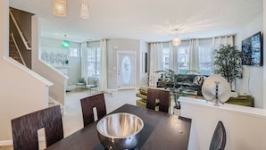 A modern living room with a dining area, featuring a staircase, large windows, and decorative lighting.