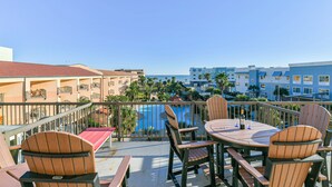 A balcony with wooden chairs and a table, overlooking a swimming pool and a blue building. - Pelican Bungalow! Top Floor, Endless Gulf Views, Pet Friendly! (Galveston)