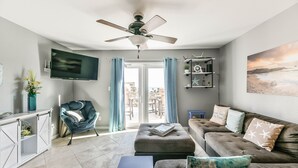 A modern living room with a flat-screen TV, a ceiling fan, a sofa with decorative pillows, a chair, and a bookshelf. - Pelican Bungalow! Top Floor, Endless Gulf Views, Pet Friendly! (Galveston)