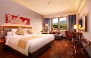 A hotel room with a large bed, a desk with a chair, a TV, and a window with a view of mountains. - Monopoli Hotel Bukittinggi (Bukittinggi)
