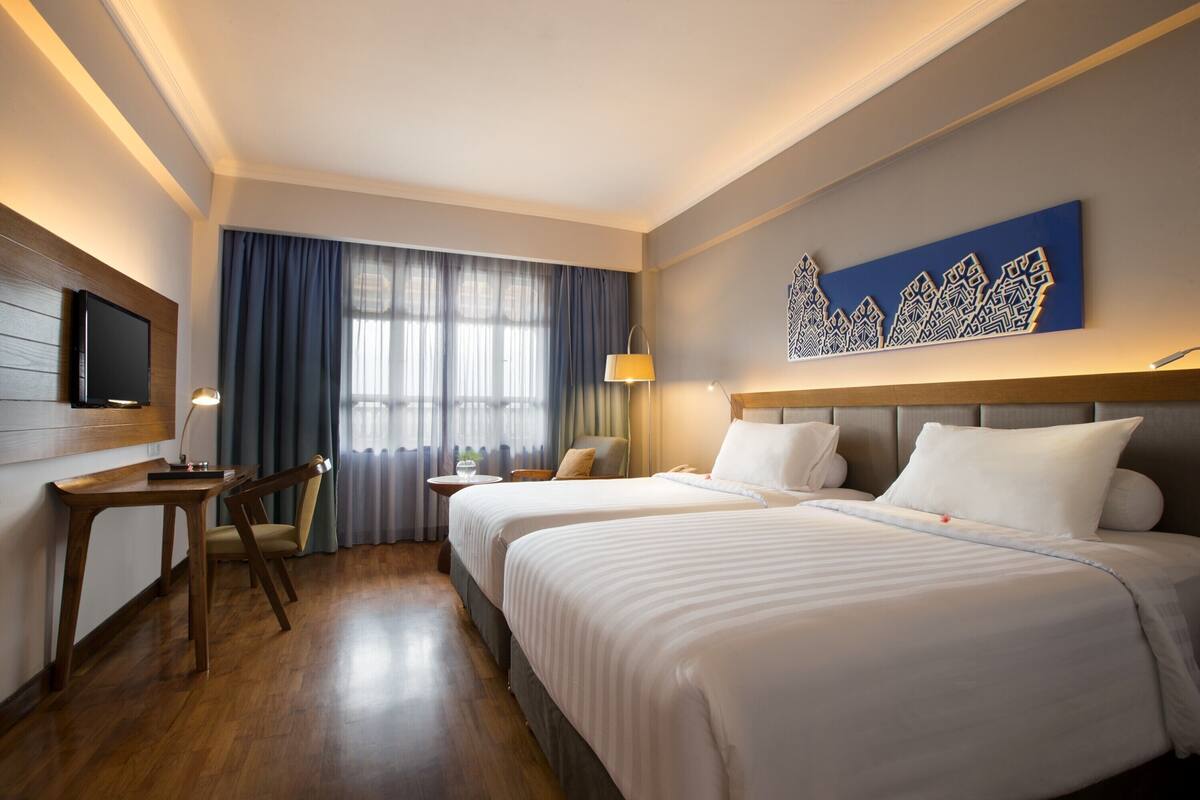 A hotel room with two beds, a desk, a chair, a TV, and a window with curtains.