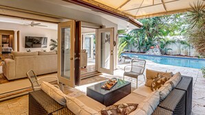 A spacious living area with a sofa, coffee table, and chairs, leading to a pool area with a patio and tropical plants. - Heated pool / Quiet Neighborhood / Family Vacation / Escape Key (Fort Lauderdale)