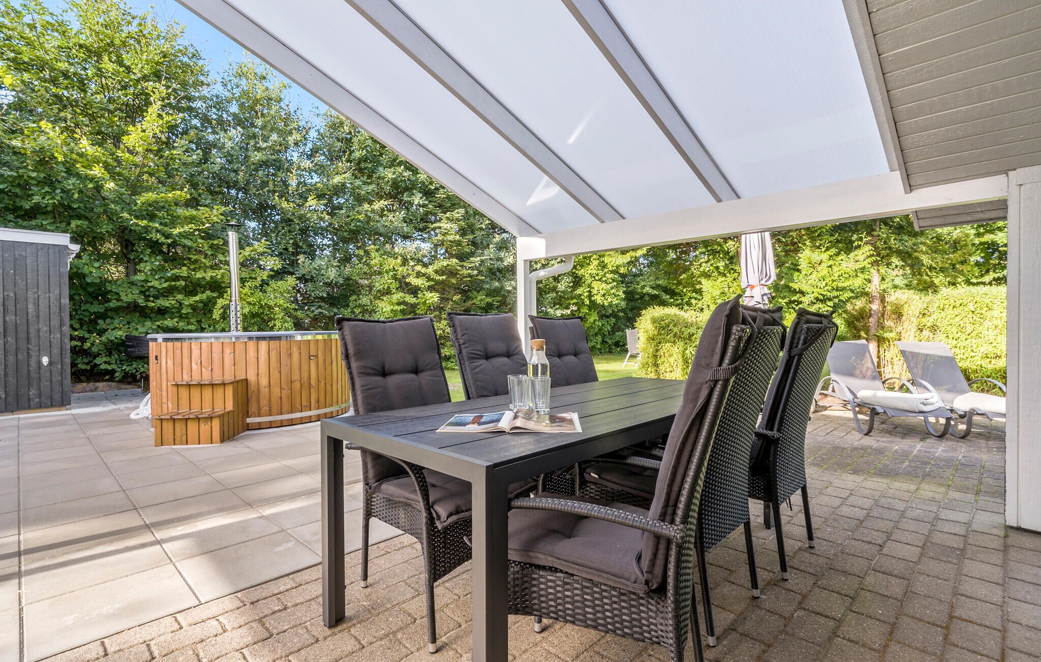 An outdoor patio with a table and chairs, a wooden hot tub, and a covered area with a view of the garden.