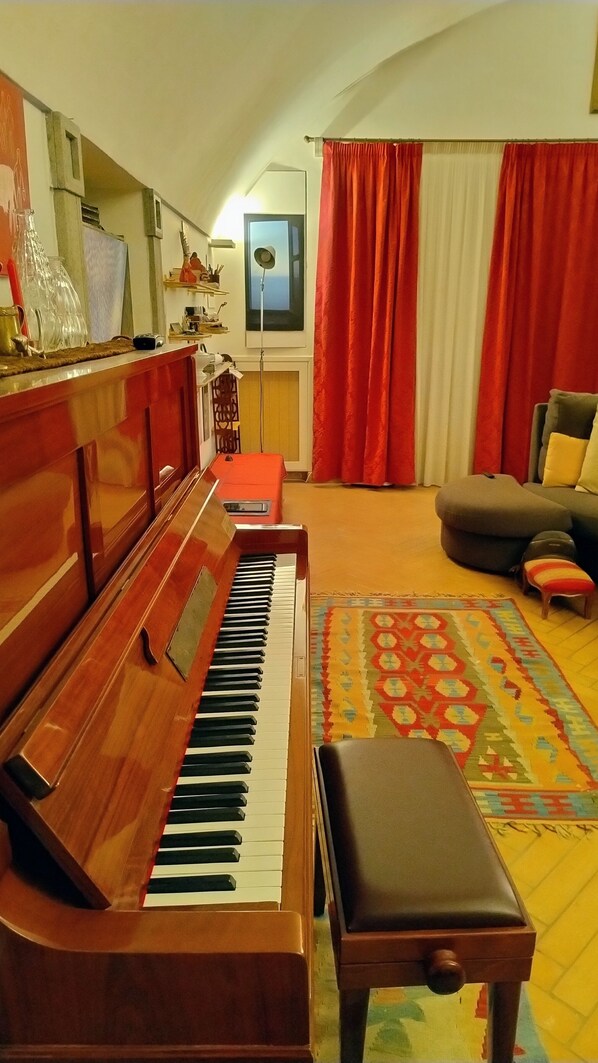 Een woonkamer met een houten piano, een klein krukje, een kleurrijk vloerkleed en een bank.