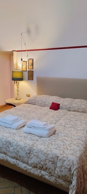 A neatly made bed with a floral-patterned bedspread, a red pillow, and folded towels on the side.