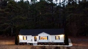 Une maison blanche à deux étages, avec un toit sombre, entourée d’une forêt.