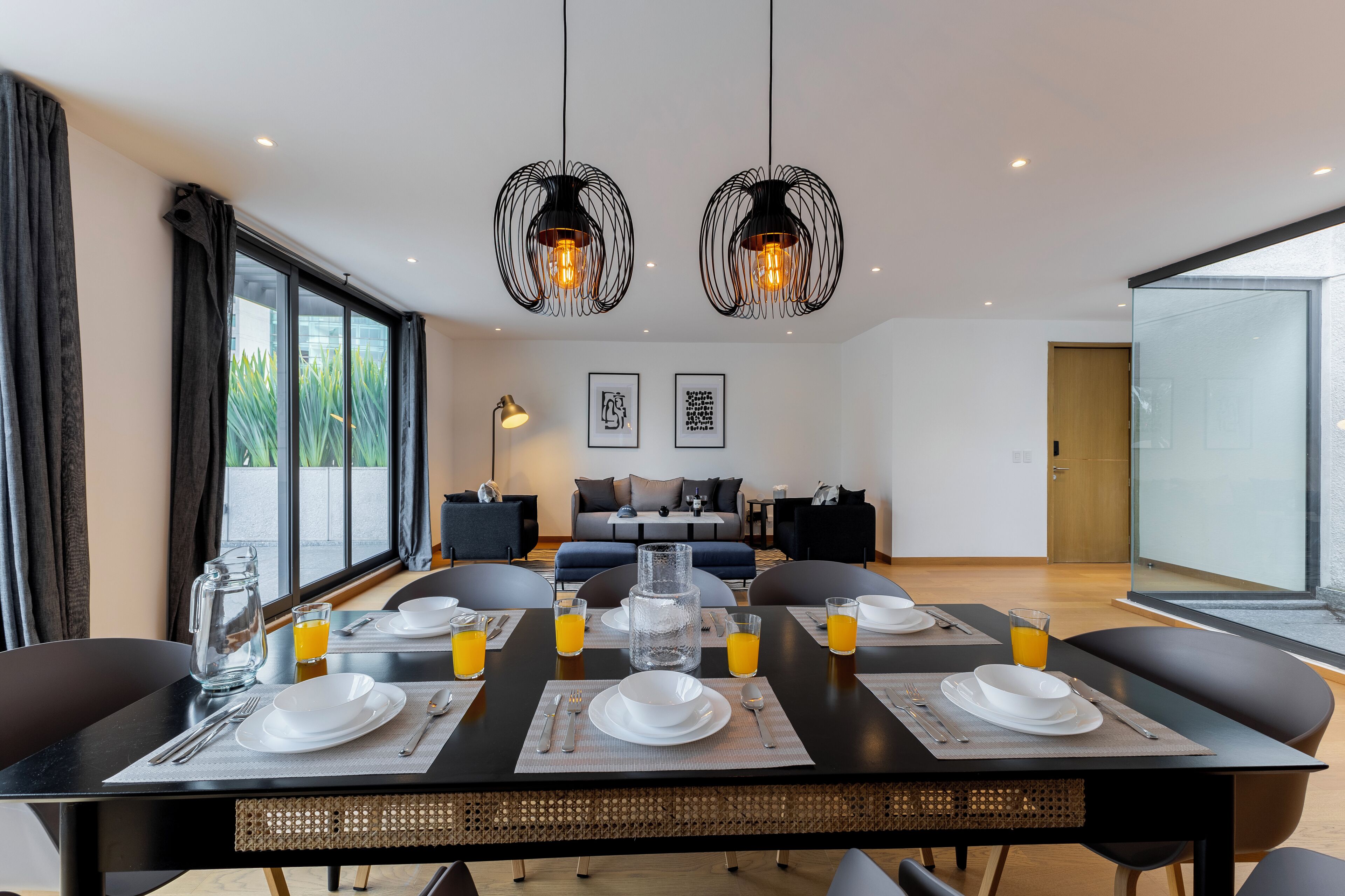 Un comedor moderno con una mesa grande preparada para el desayuno, dos lámparas colgantes y vistas al exterior a través de amplios ventanales.