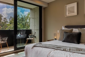 A modern bedroom with a large bed, a balcony with a glass door, and a view of greenery. - Capitalia Galileo Residences (Mexico City)