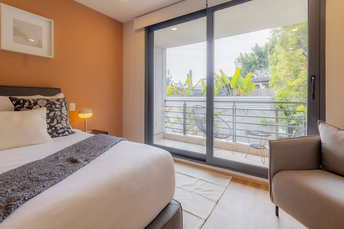 A hotel room with a large bed, a sofa, a balcony with a view of trees and a railing.