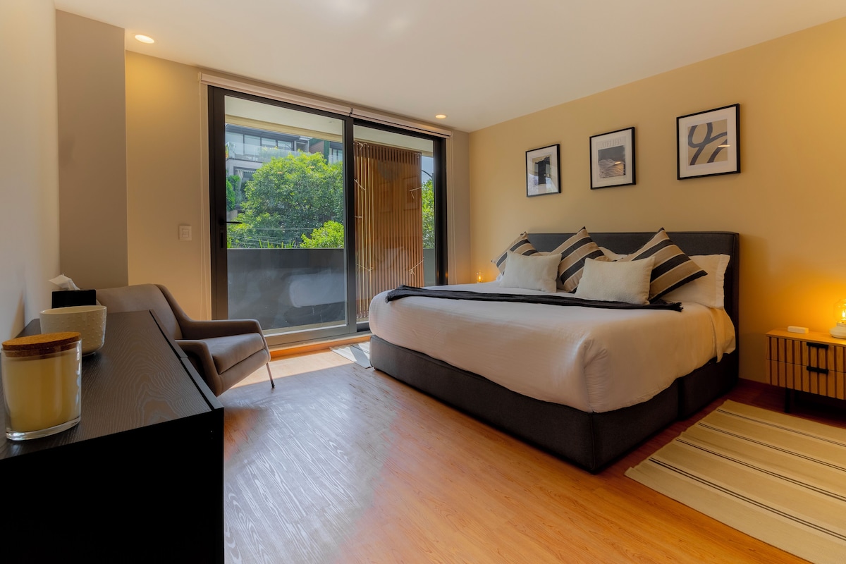 A modern bedroom with a large bed, a desk, and a chair. There is a sliding glass door leading to a balcony with a view of greenery.