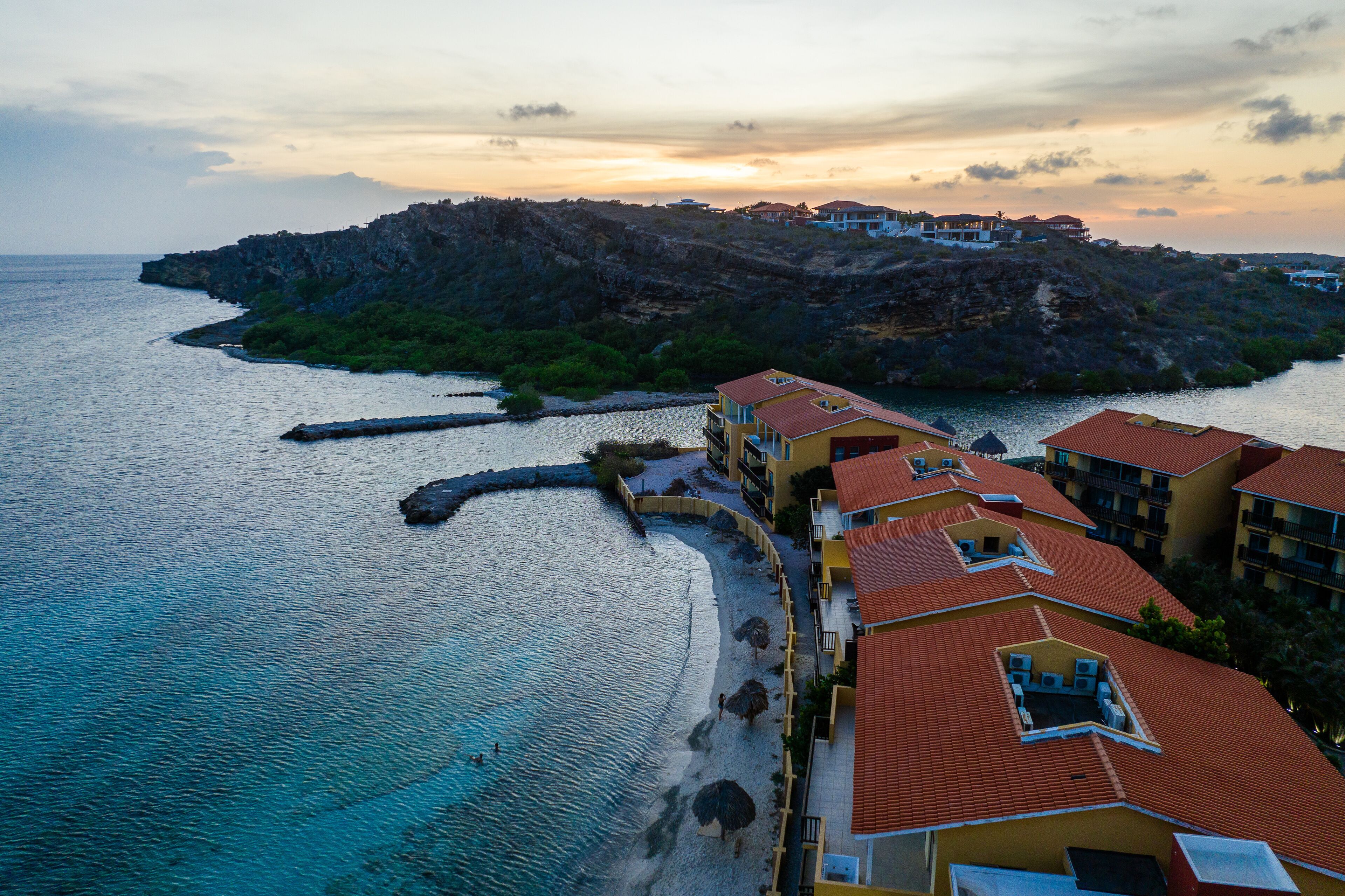 Ein Küstenhotel mit mehreren Balkonen mit Blick auf das Meer, eine felsige Insel im Hintergrund und ein klarer Himmel.