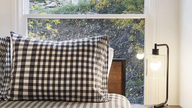 Une chambre à coucher avec une couvre-lit à carreaux, une lampe de chevet et une fenêtre donnant sur un paysage verdoyant et un bâtiment.