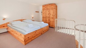 Una cama de madera con dos almohadas y una manta blanca, un armario de madera, una cuna blanca y una alfombra de tonos claros.