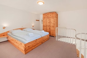 Un lit en bois avec deux oreillers et une couverture blanche, une armoire en bois, un berceau blanc et un tapis de couleur claire.