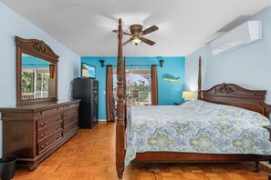 A bedroom with a wooden four-poster bed, a ceiling fan, a dresser, and a large window with a view of the outdoors.