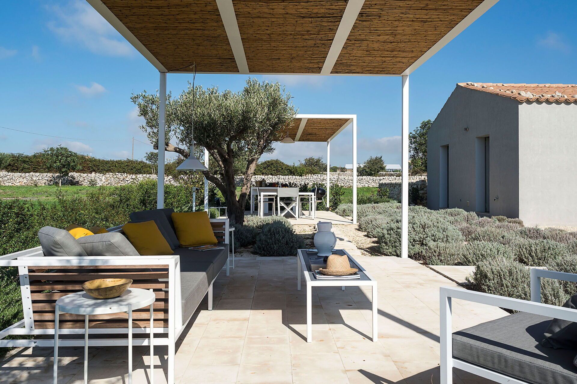 An outdoor patio with a wooden pergola, white furniture, and a view of a stone wall and greenery in the background.