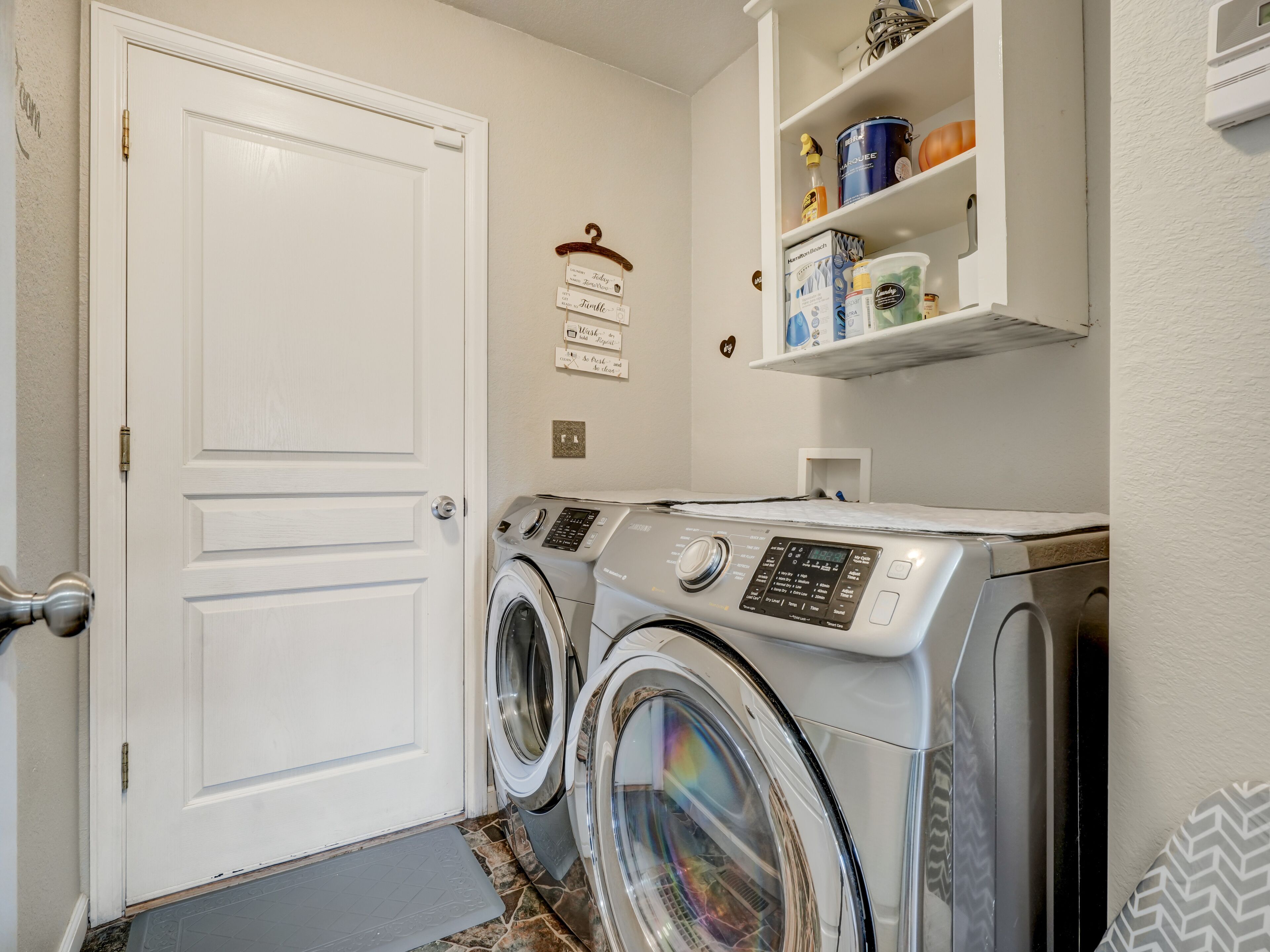 A laundry room with two front-loading washing machines, a white door, a shelf with cleaning supplies, and a small step leading to the door.
