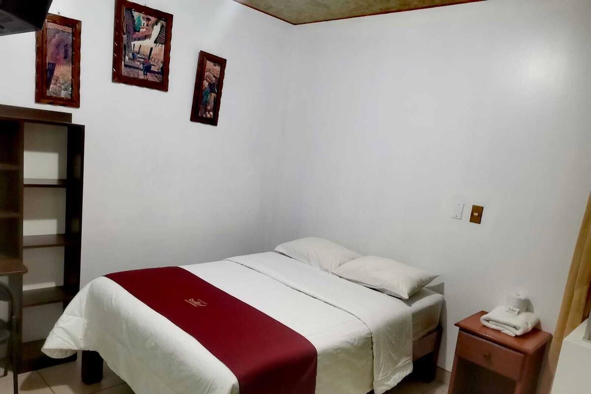 Une chambre avec un lit simple, une couvre-lit rouge et blanche, une table de chevet en bois et une petite étagère.