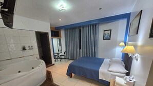 A hotel room with a large bathtub, a bed with a blue bedspread, a bedside table with a lamp, and a small desk with a chair. - HOTEL LOS ANGELES (Huaral)