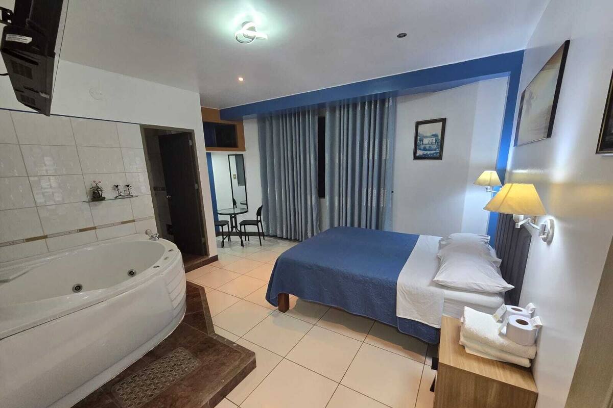 Une chambre d’hôtel avec une grande baignoire, un lit recouvert d’une courtepointe bleue, une table de chevet avec une lampe et un petit bureau avec une chaise.