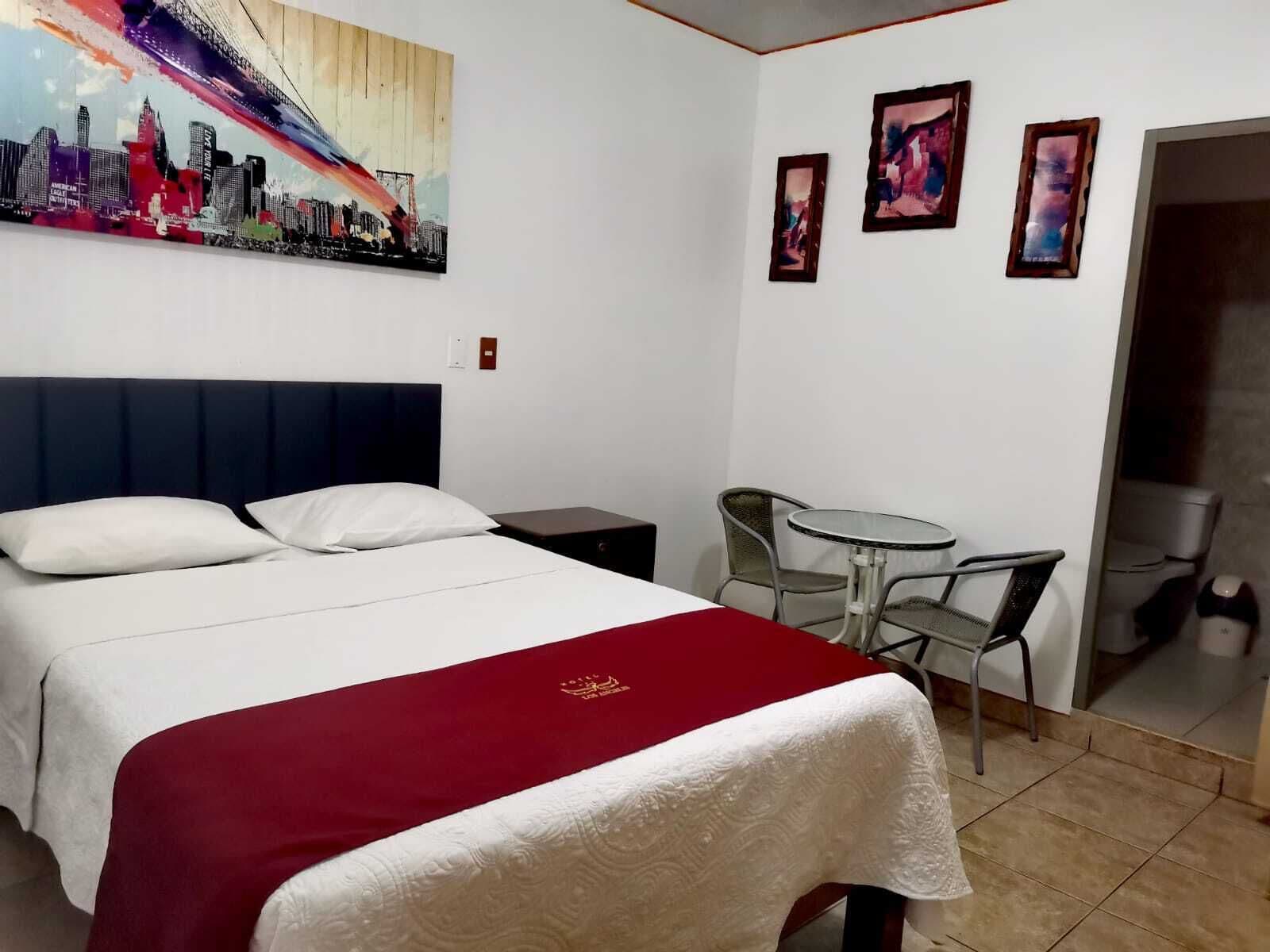 A hotel room with a bed, a small table with chairs, a painting on the wall, and a door leading to a bathroom.