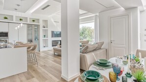 A modern kitchen with a central island, a dining area with a table set for four, and a living room with a sofa and chairs.