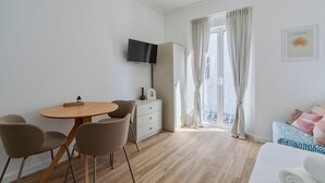 Et moderne hotelværelse med et træspisebord, to stole, et fladskærms-TV, en hvid kommode, en balkon med gardiner og en seng med mønstret sengetøj.