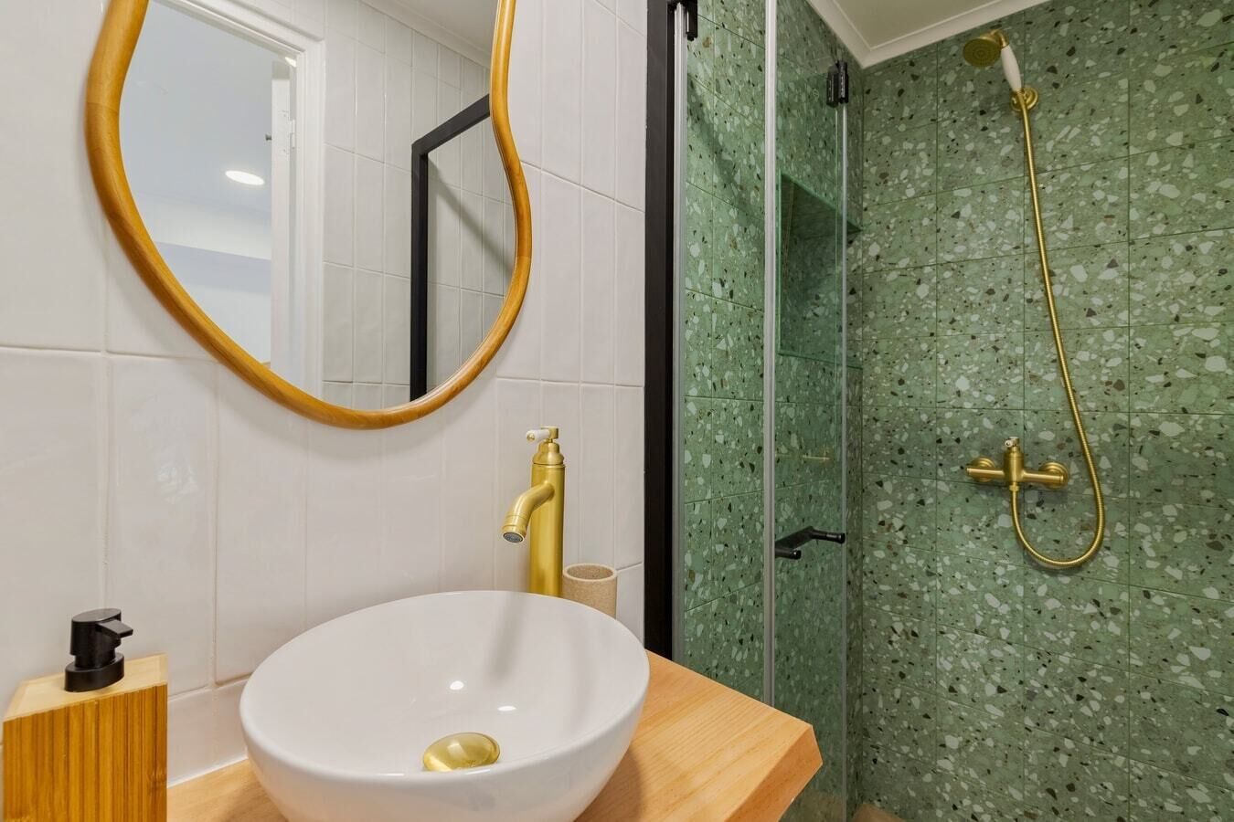Um banheiro com pia branca, torneiras douradas, penteadeira de madeira, espelho redondo e chuveiro com azulejos verdes com manchas.
