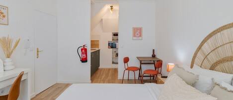 Ein Schlafzimmer mit einem Bett, einem Schreibtisch und einem Stuhl. An der Wand hängt ein Feuerlöscher und es steht ein kleiner Tisch mit Stühlen bereit.