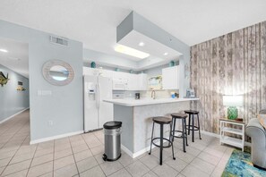A modern kitchen with white cabinets, a central island with stools, a trash bin, and a wooden accent wall.