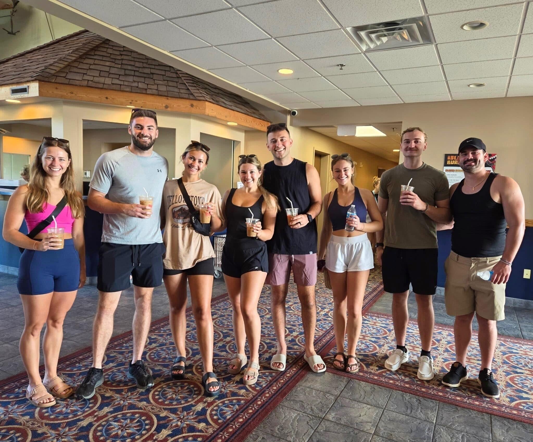 A group of people standing in a hotel lobby with a patterned carpet, a ceiling with recessed lighting, and a visible wall with a textured finish.