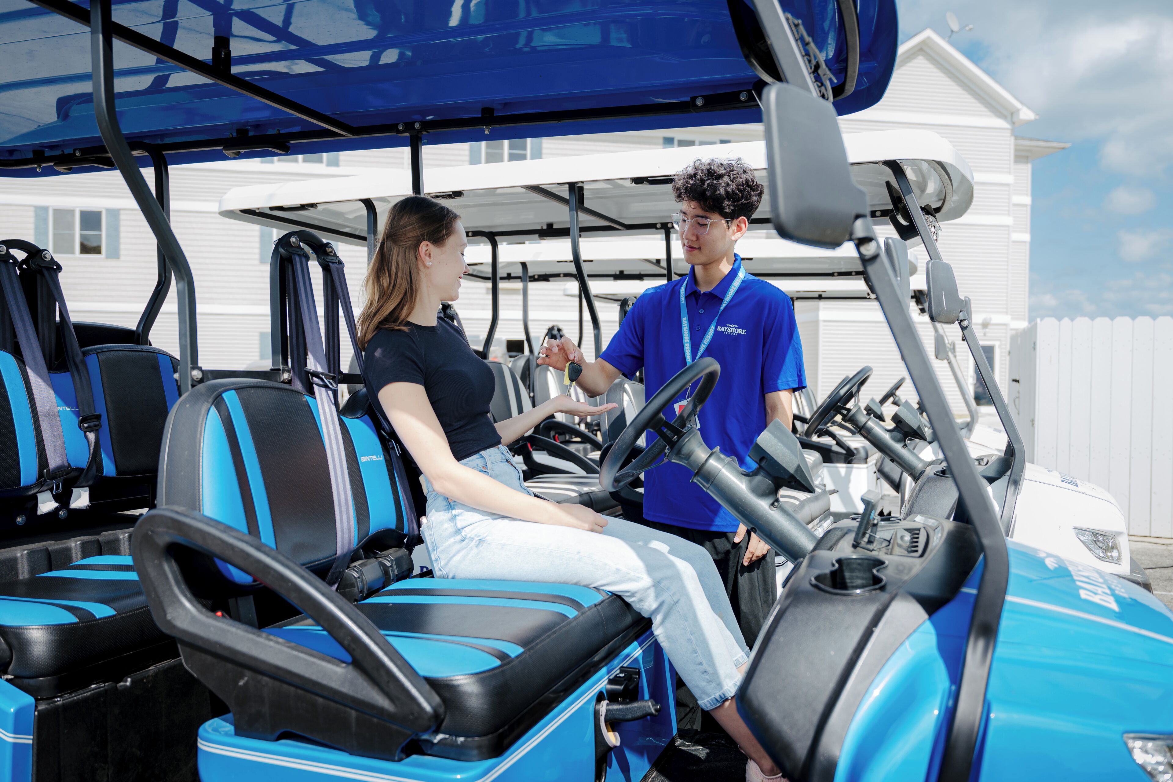 A golf cart with blue seats and a steering wheel, a person in a blue shirt standing beside it, and a white building in the background.
