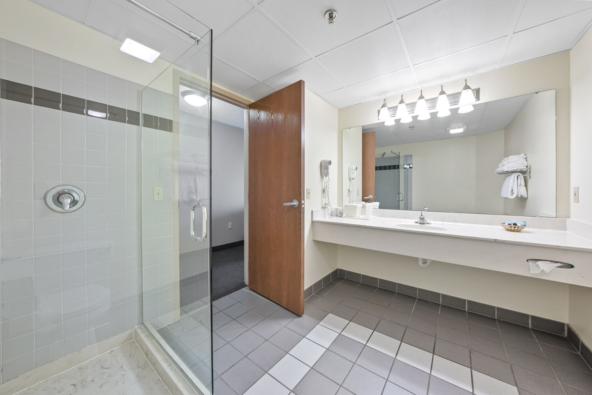 A modern bathroom with a glass shower enclosure, a wooden door, a large mirror, and a white sink with a towel rack.